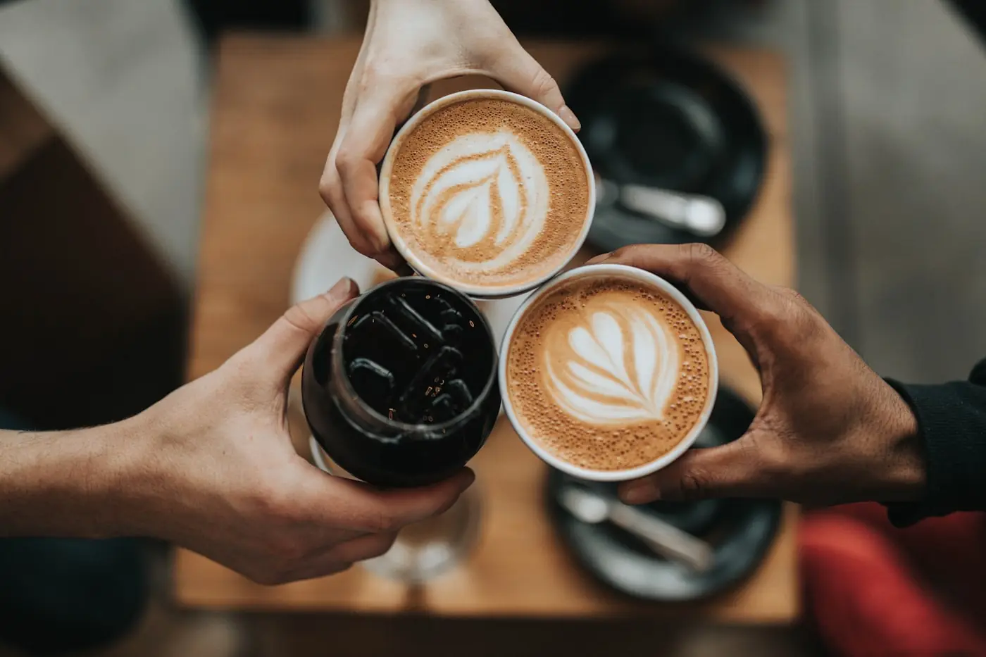 Beberapa tangan mengangkat cangkir kopi bersulang di atas meja kayu.